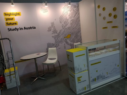 Eine Großaufnahme von dem "highlight your futur - study in Austria" Stand, welcher aus einem Tresen im Vordergrund und einem kleinen Tisch mit Sessel links dahinter besteht.