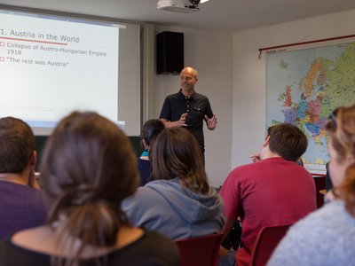 Ein Vortragender spricht zu Studierenden in einem Hörsaal mit eine Landkarte an der Wand. . 