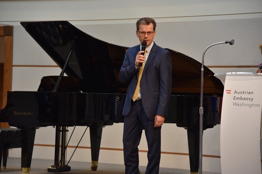 Clemens Mantl with a microphone stands on a stage with a piano behind him