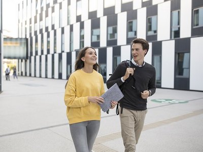 Eine Studentin und ein Student gehen nebeneinander vor einem Gebäude. 