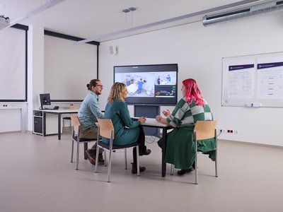 Drei Personen sitzen an einem Tisch vor einem digitalen Bildschirm.