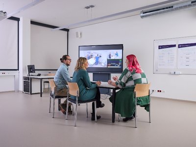 Drei Personen sitzen um einen Tisch in einem Unterrichtsraum.