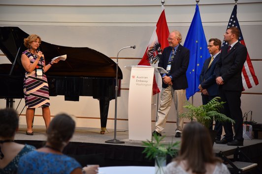 A woman moderates on a stage, while three men stand behind a podium and in front of the Austrian, American and EU flags.