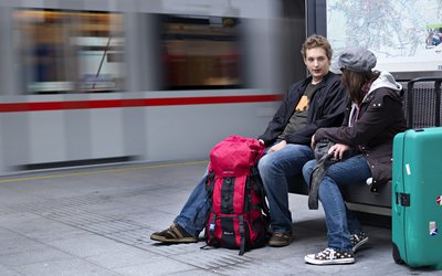Zwei jugendliche Burschen sitzen auf einer Bank an einem Bahnsteig mit Rucksack und Koffer, im Hintergrund ein fahrender Zug.