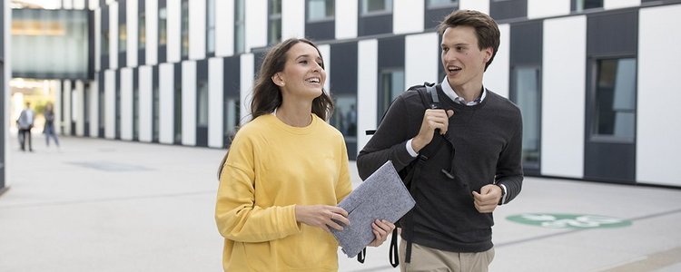 Eine Studentin und ein Student gehen nebeneinander vor einem Gebäude. 