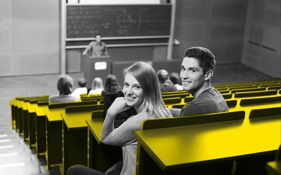 Eine Studentin und ein Student sitzen im Hörsaal und lachen in die Kamera. 